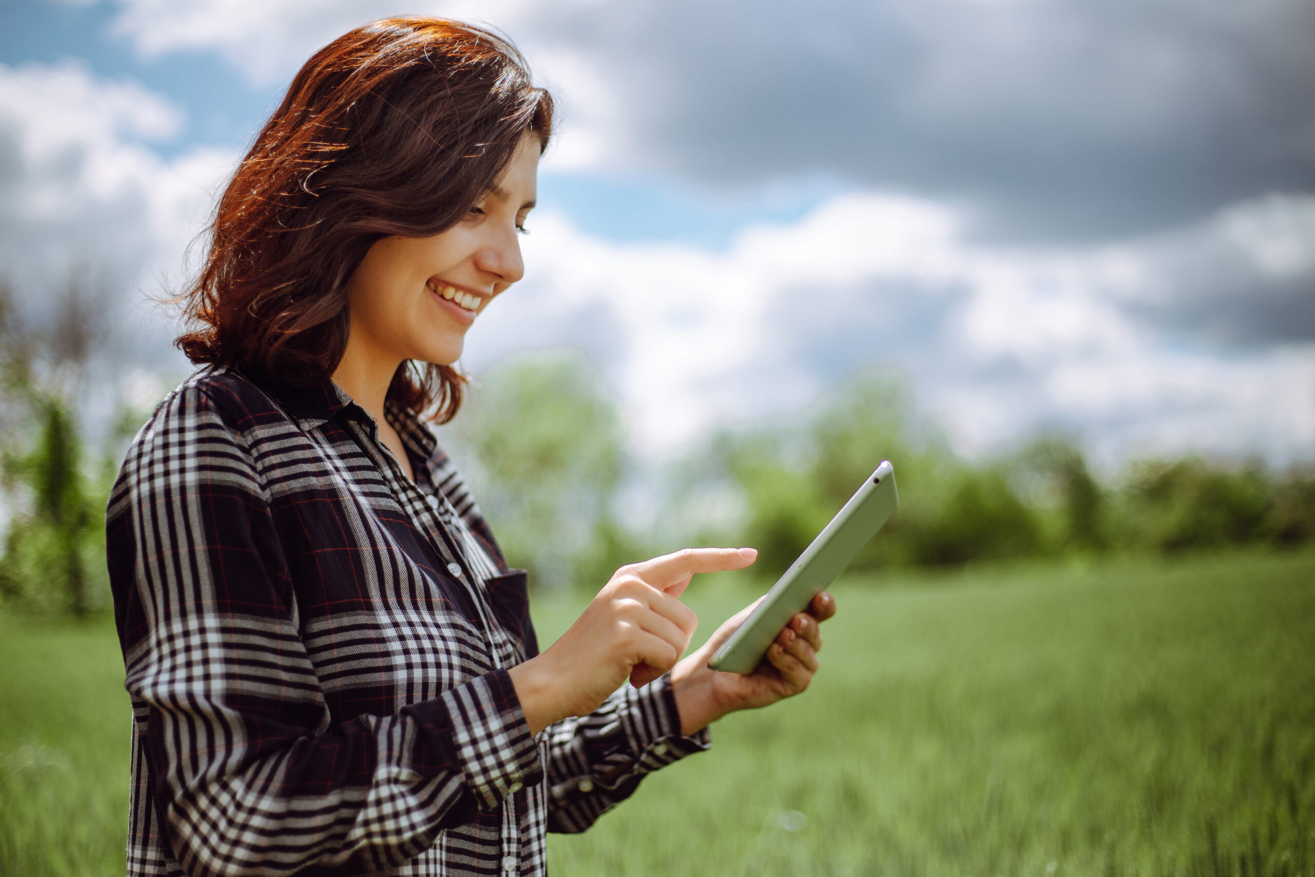 Young woman farmer wearing checkered shirt is checking harvest progress on a tablet at the green wheat field. New crop of wheat is growing. Agricultural and farm concept