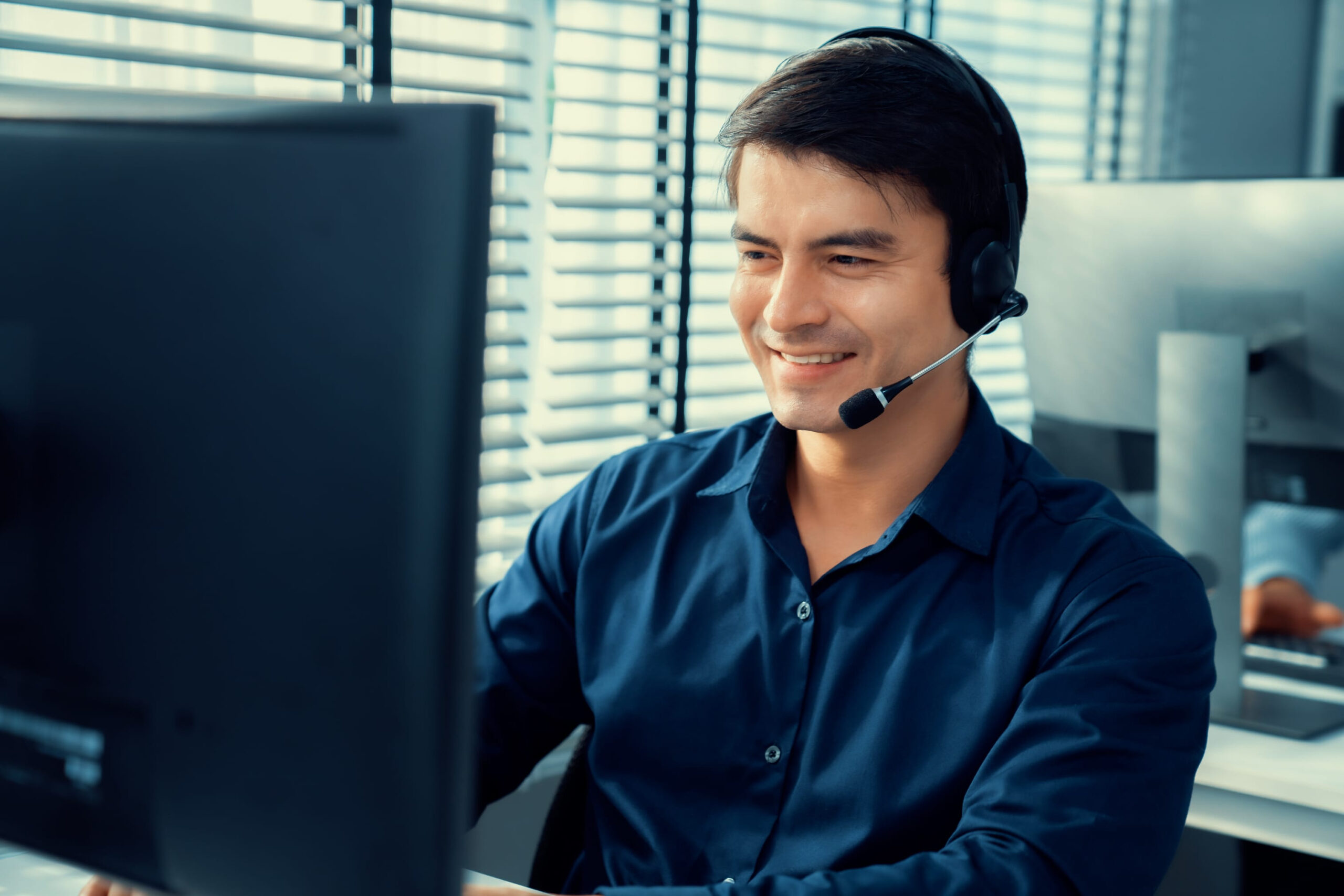 Young competent asian male call center agent working at his computer while simultaneously speaking with customers. Concept of an operator, customer service agent working in the office with headset.